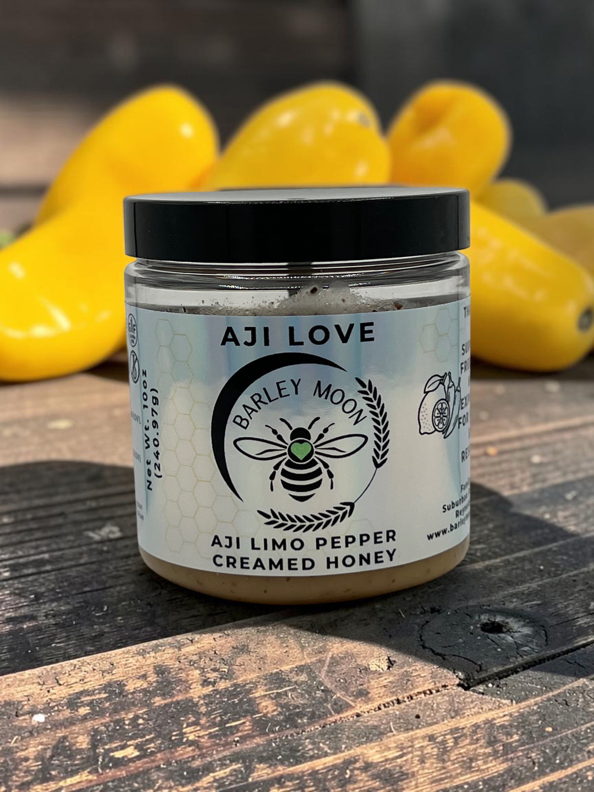 Barley Moon Botanicals Aji Limo Creamed Honey jar with a black lid, sitting on a wooden surface with yellow peppers.