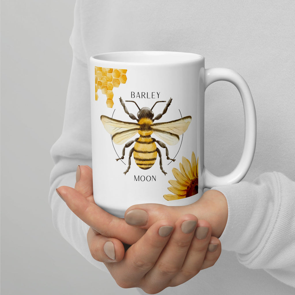 Barley Moon Botanicals 15 oz white ceramic mug featuring a bee, honeycomb, and flower illustration held in hands.