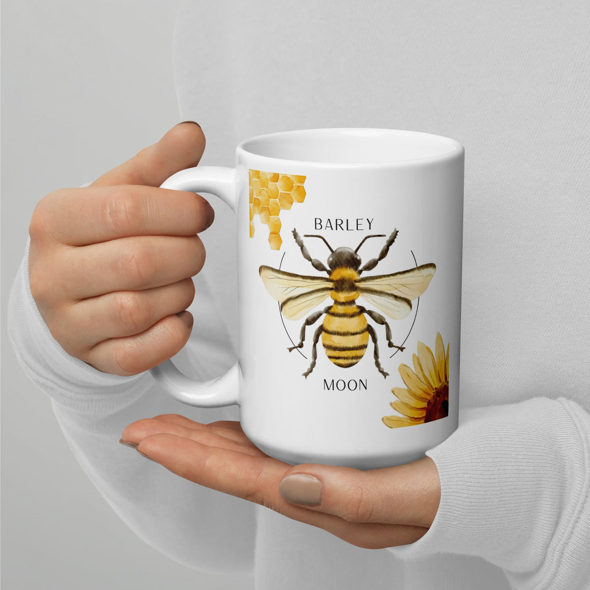 Barley Moon Botanicals 15 oz white ceramic mug with a bee, honeycomb, and flower illustration held in two hands.