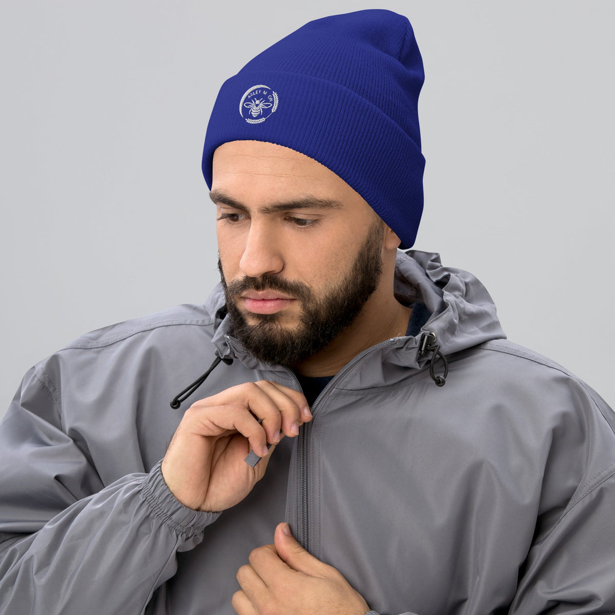 A man wearing a blue Barley Moon Botanicals cuffed beanie with a white bee and moon logo, paired with a grey jacket.