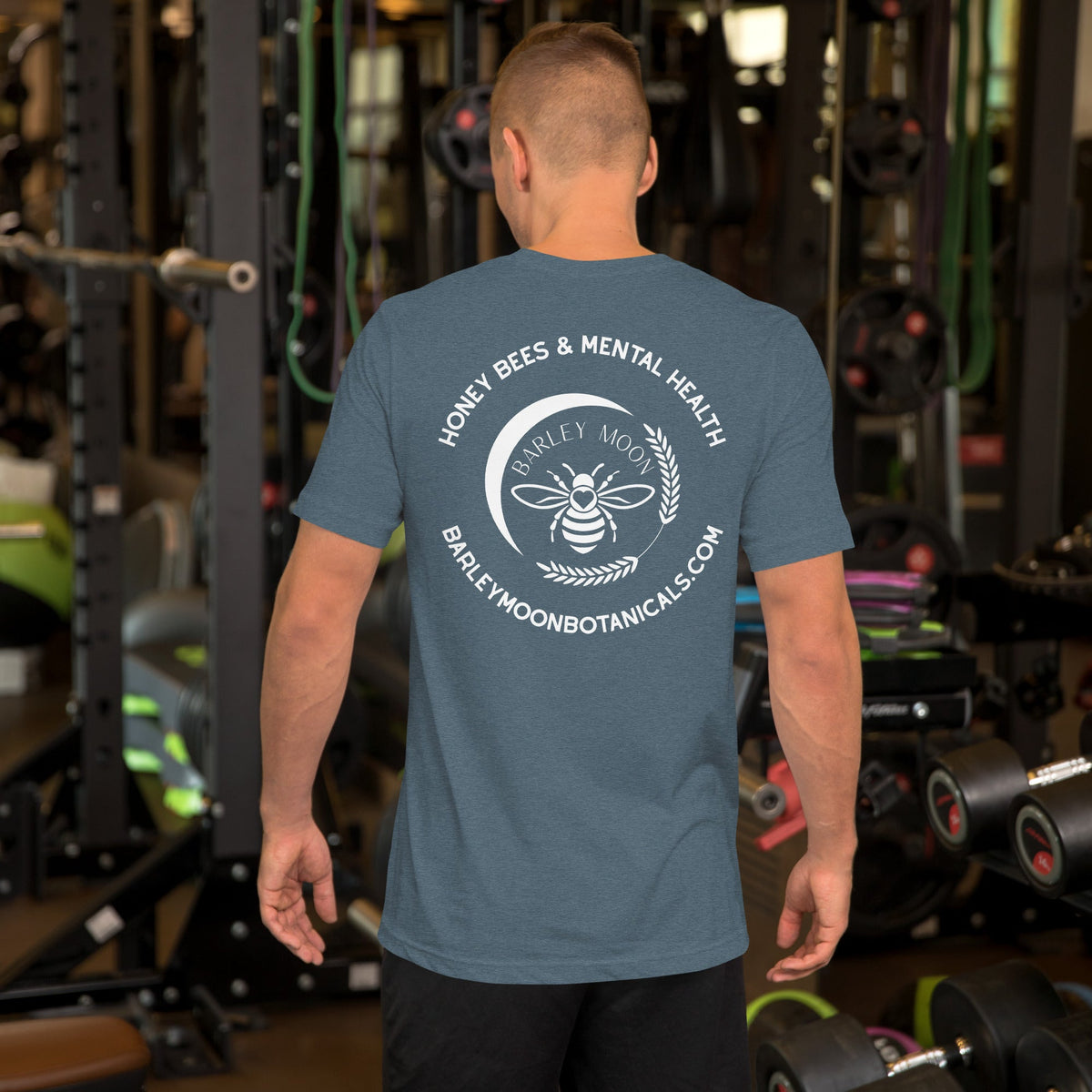 A man wearing a blue Barley Moon Botanicals t-shirt with a white bee and crescent moon logo on the back in a gym.