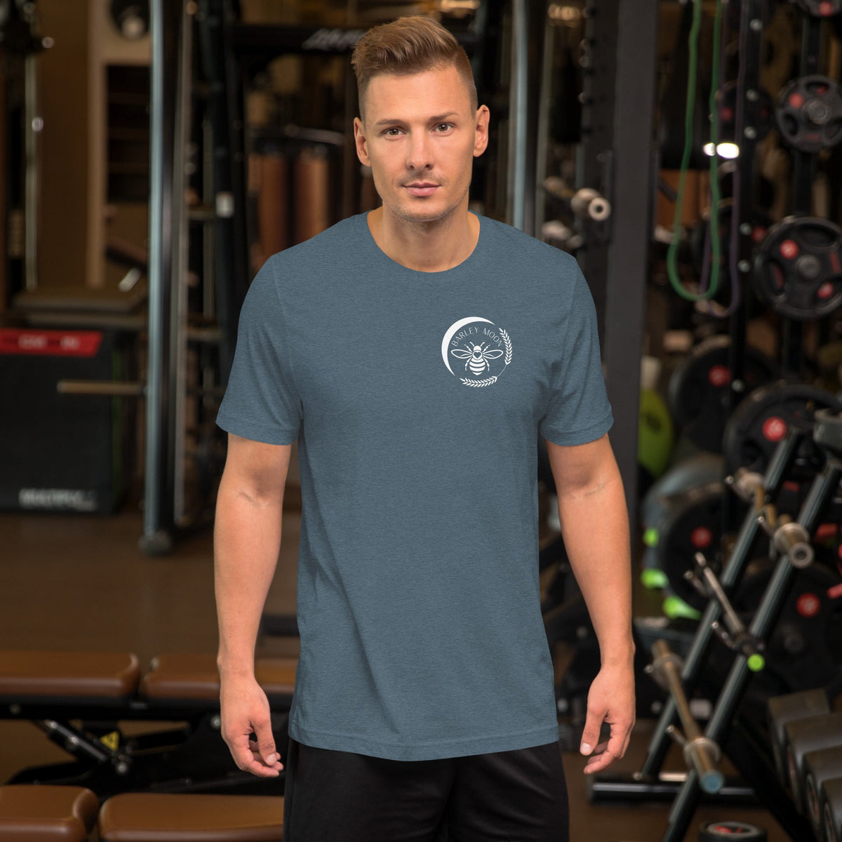 A man wearing a blue Barley Moon Botanicals unisex t-shirt with a white bee logo in a gym setting.