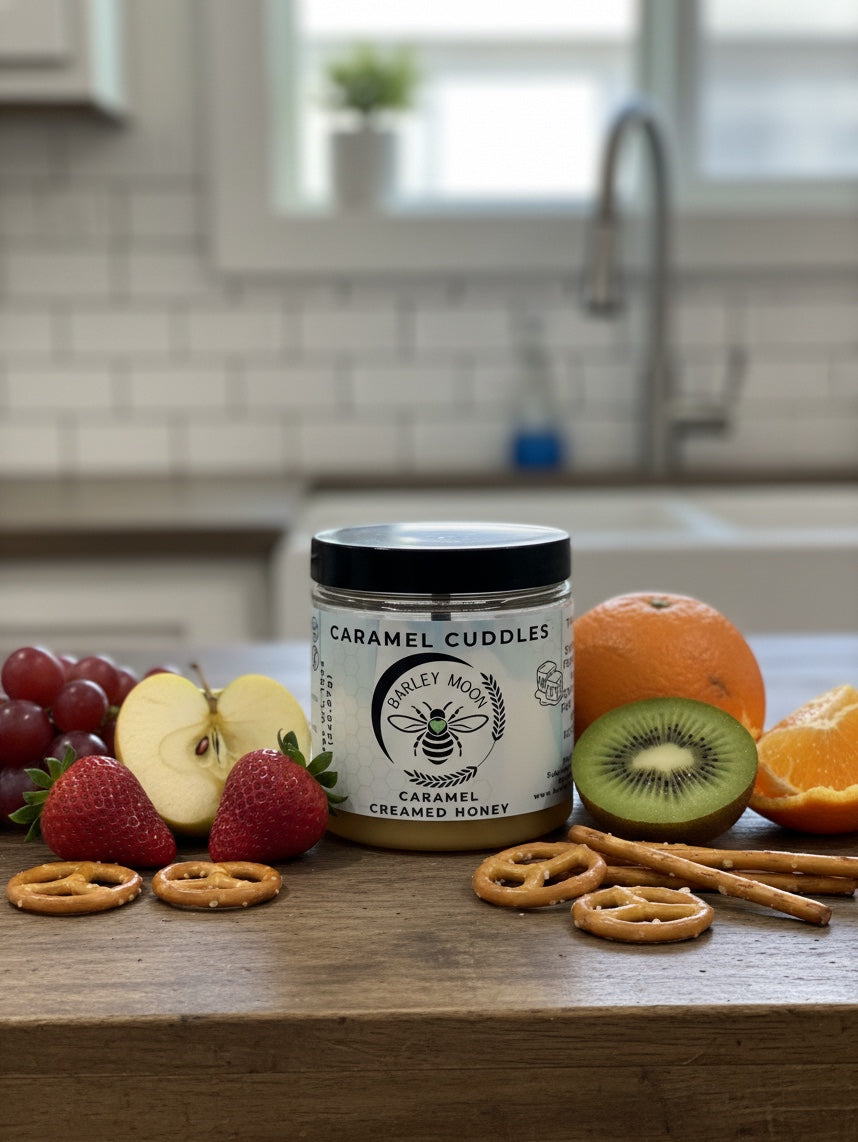 Barley Moon Botanicals Caramel Cuddles creamed honey jar surrounded by fresh fruit and pretzels on a wooden table.