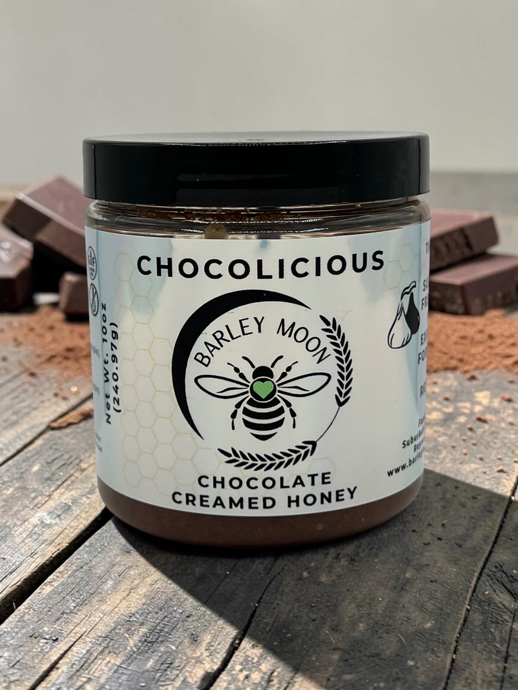 Barley Moon Botanicals Chocolicious chocolate creamed honey jar on a rustic wooden surface with cocoa powder and chunks.