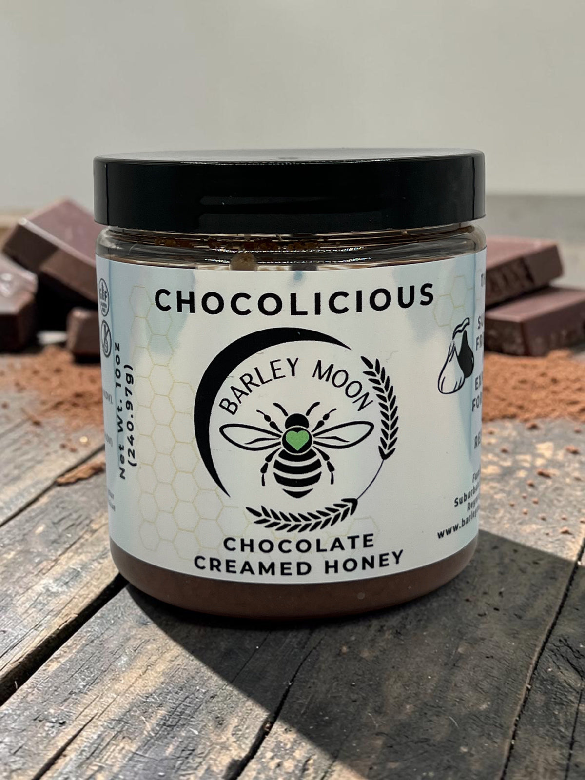 Barley Moon Botanicals Chocolicious chocolate creamed honey jar on a rustic wooden surface with cocoa powder and chunks.