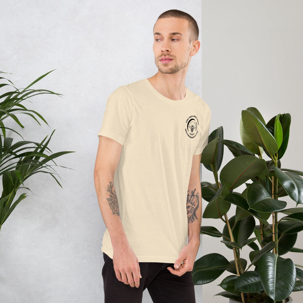 A man wearing a Barley Moon Botanicals cream unisex t-shirt with a small bee logo, standing by indoor plants.