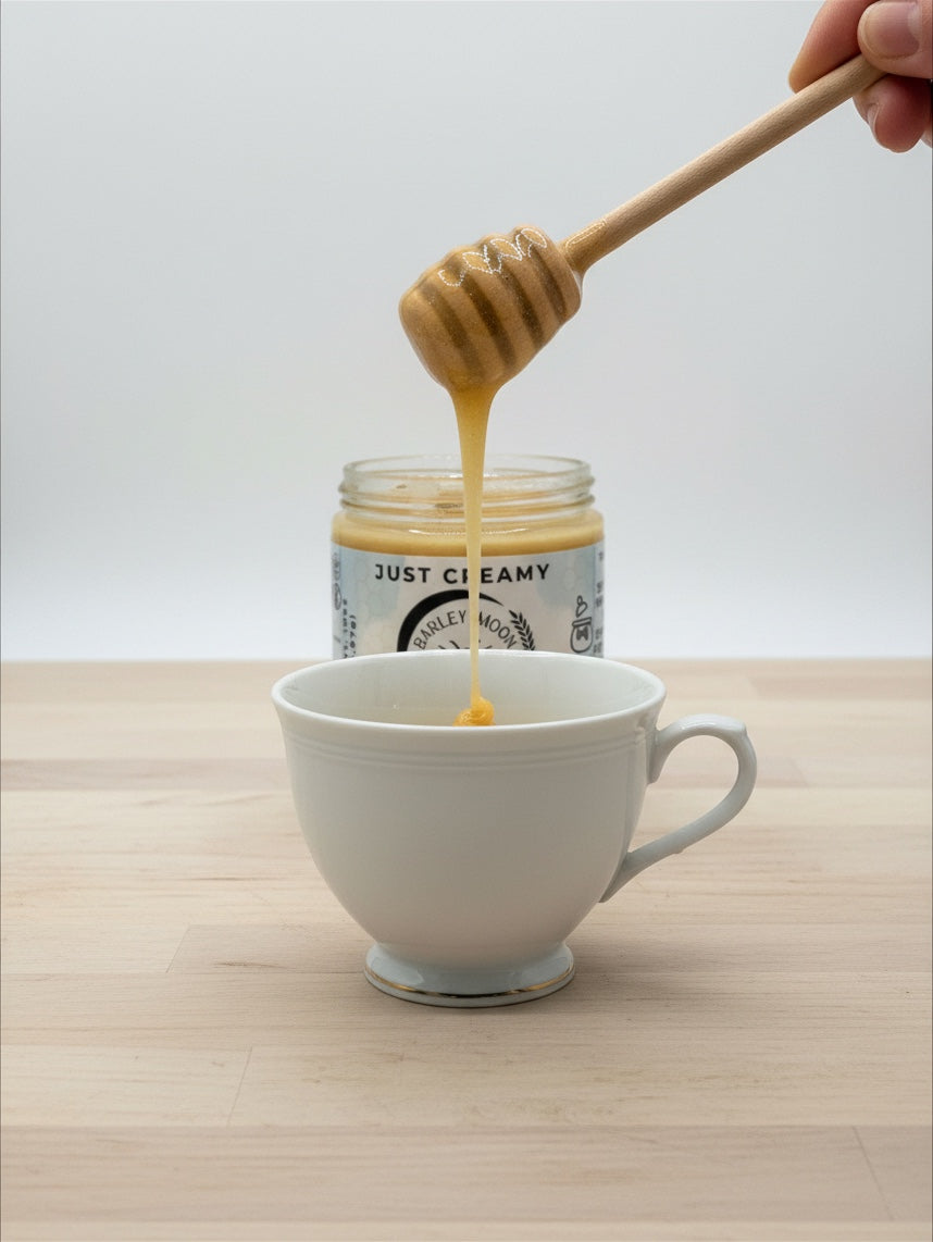 Barley Moon Botanicals Just Creamy original creamed honey drizzling from a wooden dipper into a white ceramic cup