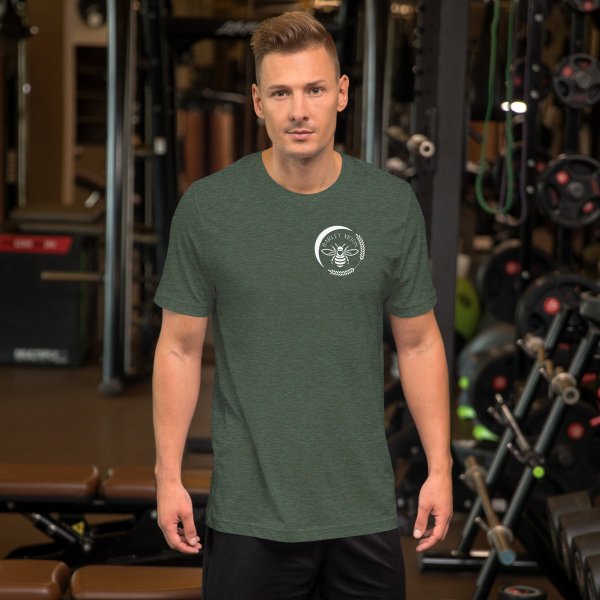 Man wearing a green Barley Moon Botanicals t-shirt with a white bee and crescent moon logo in a gym setting.