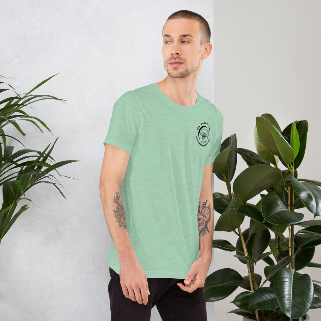 A man wearing a soft green Barley Moon Botanicals unisex t-shirt with a small bee logo on the chest, standing by plants.
