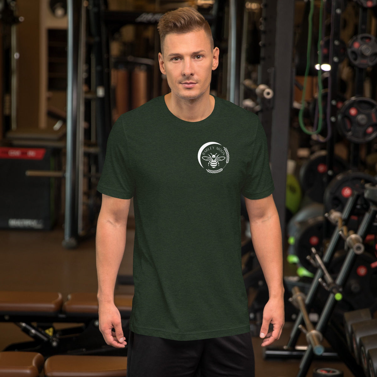 A man wearing a dark green Barley Moon Botanicals unisex cotton t-shirt with a white bee logo in a gym setting.