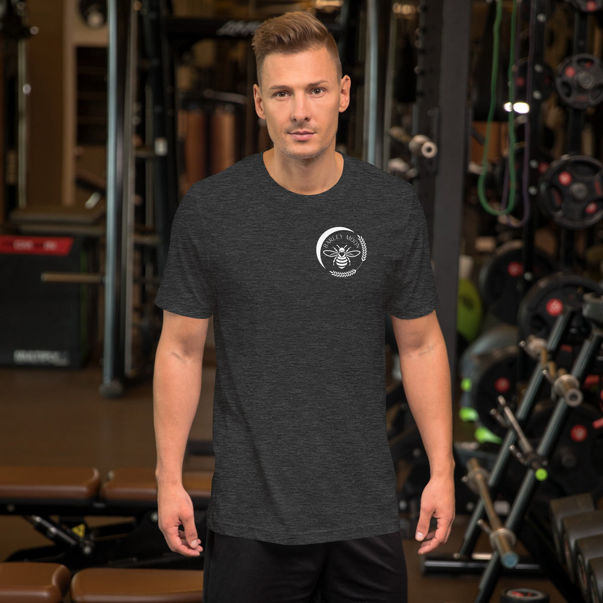 A man wearing a dark grey Barley Moon Botanicals unisex t-shirt with a white bee logo in a gym setting.