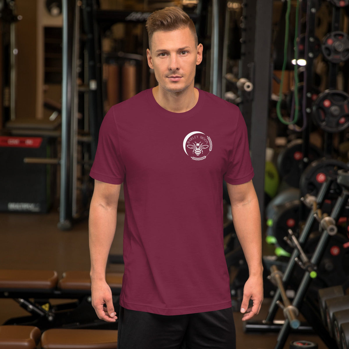 A man wearing a maroon Barley Moon Botanicals t-shirt with a white bee and crescent moon logo in a gym setting.