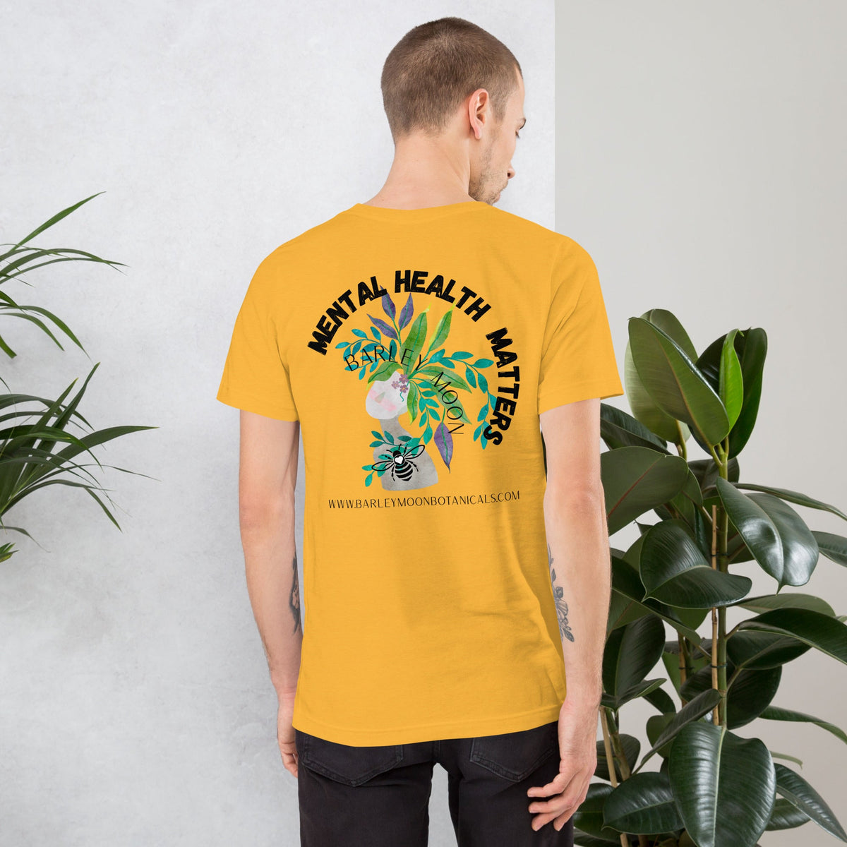 Man wearing a mustard yellow Barley Moon Botanicals unisex t-shirt with a botanical mental health graphic on the back.