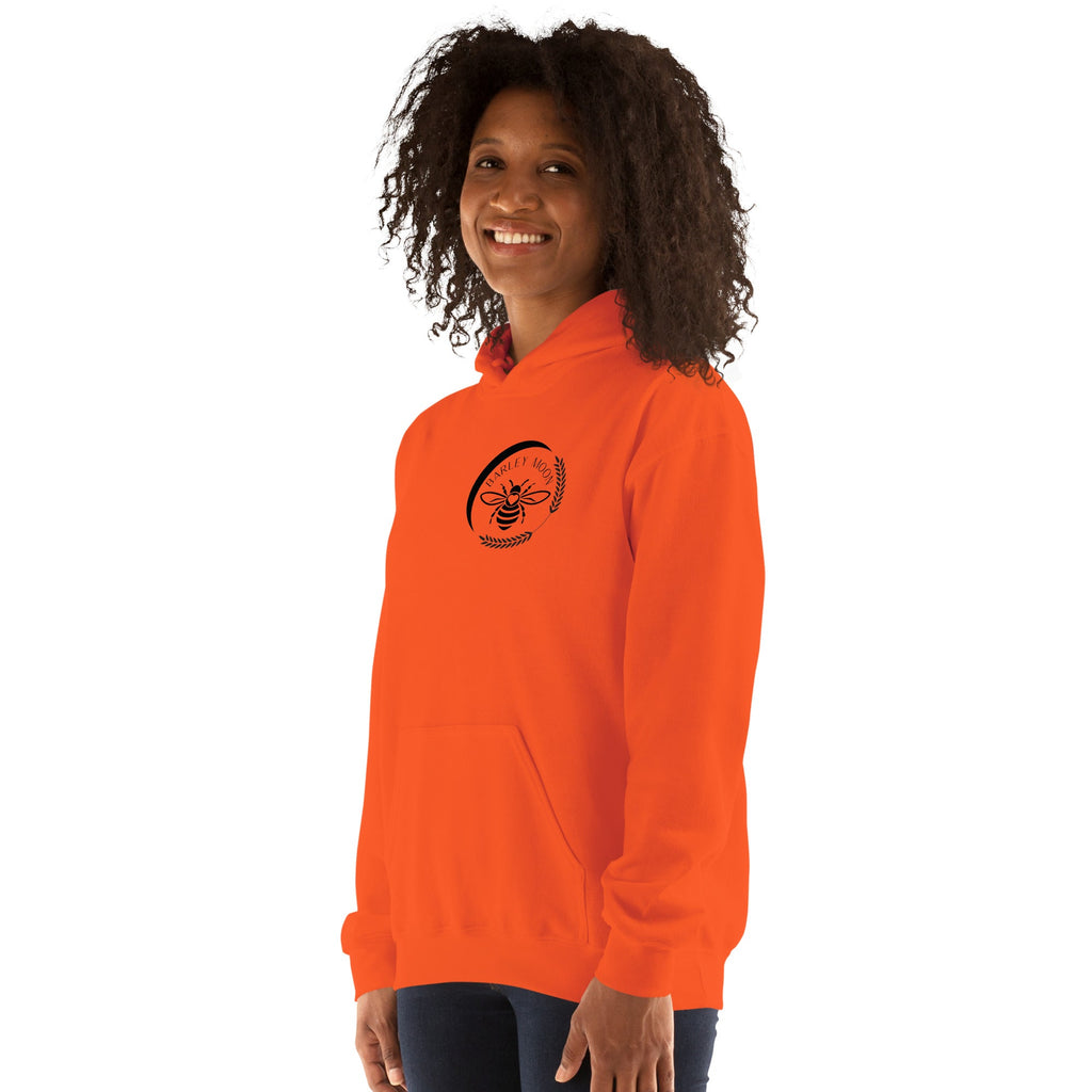 A woman wearing a bright orange Barley Moon Botanicals unisex hoodie with a black bee and crescent moon logo.