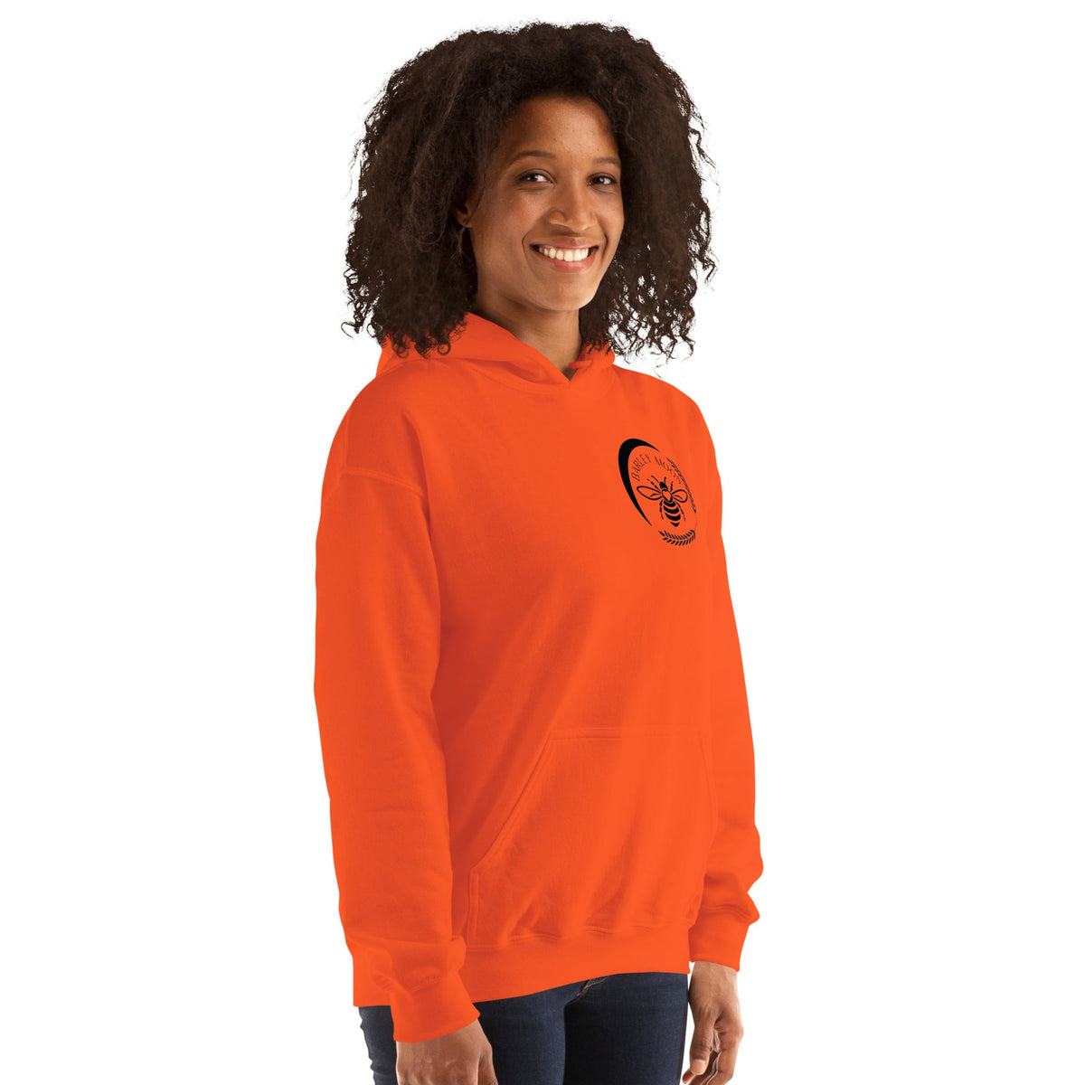 A woman wearing a bright orange Barley Moon Botanicals unisex hoodie with a black bee and crescent moon logo.