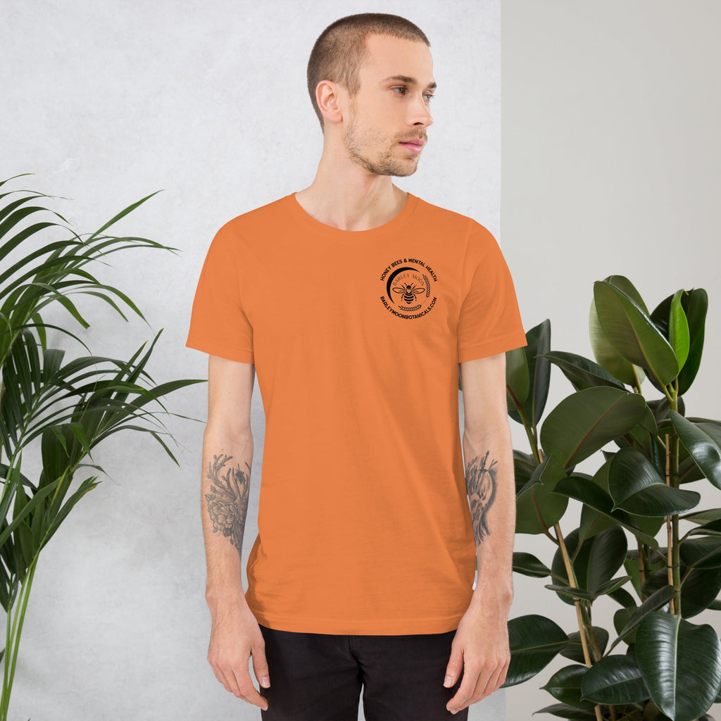 A man wearing an orange Barley Moon Botanicals unisex t-shirt with a black bee logo on the chest in a studio setting.