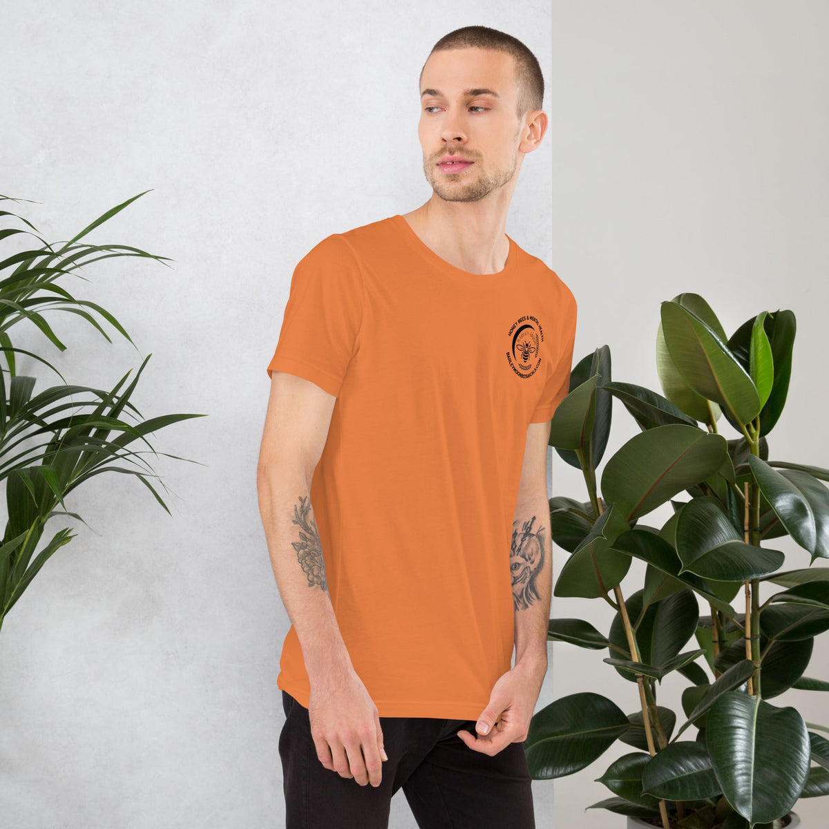 A man wearing an orange Barley Moon Botanicals unisex t-shirt with a small black bee logo on the chest.