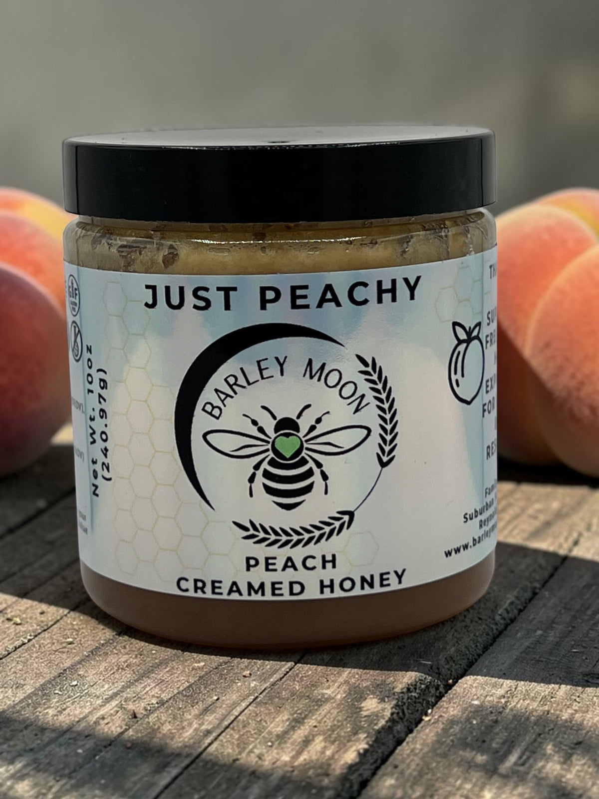 Barley Moon Botanicals Just Peachy creamed honey jar with a black lid, displayed with fresh peaches on a wooden table.