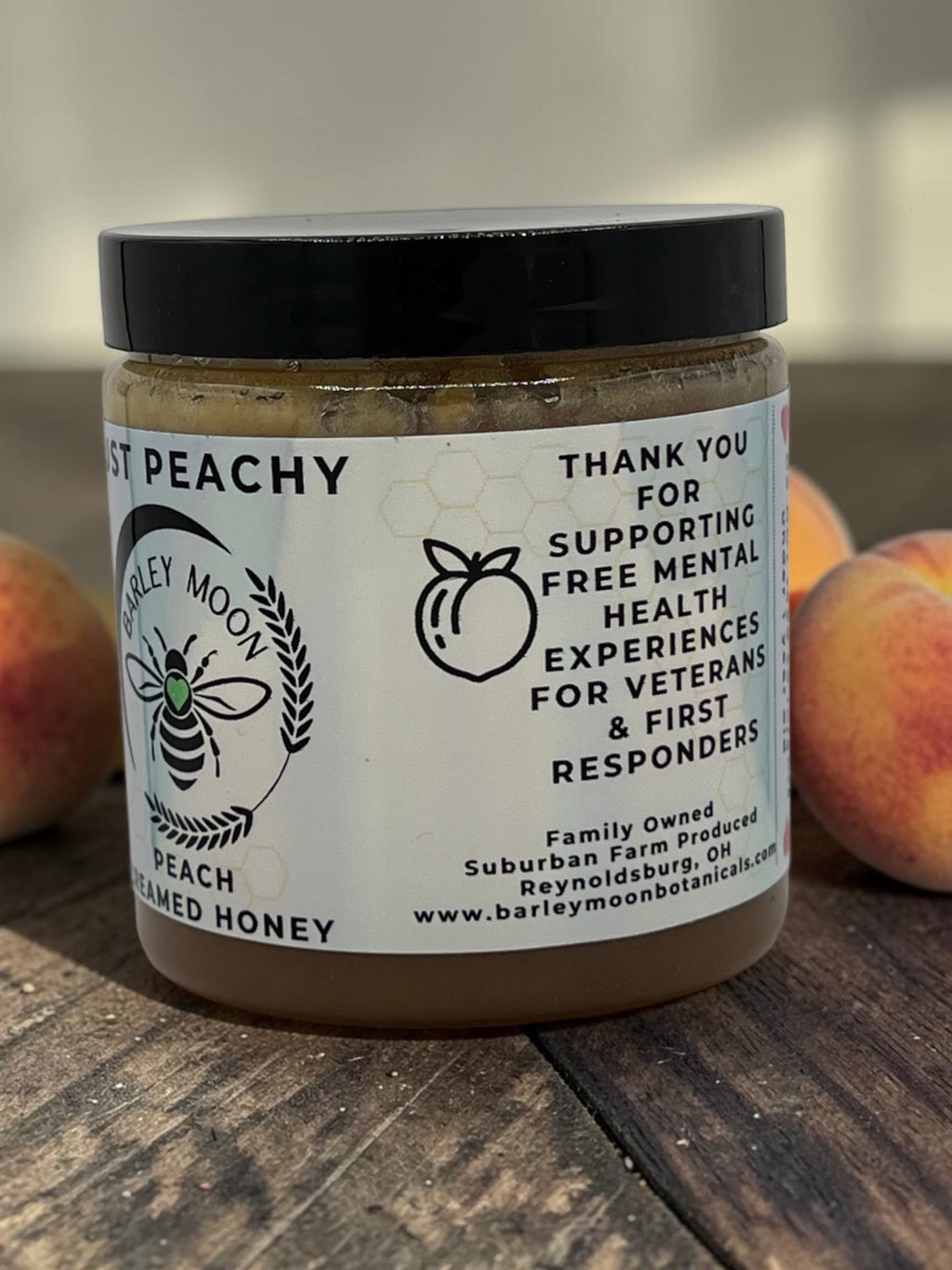 Barley Moon Botanicals Just Peachy creamed honey jar on a wooden surface with fresh peaches in the background.
