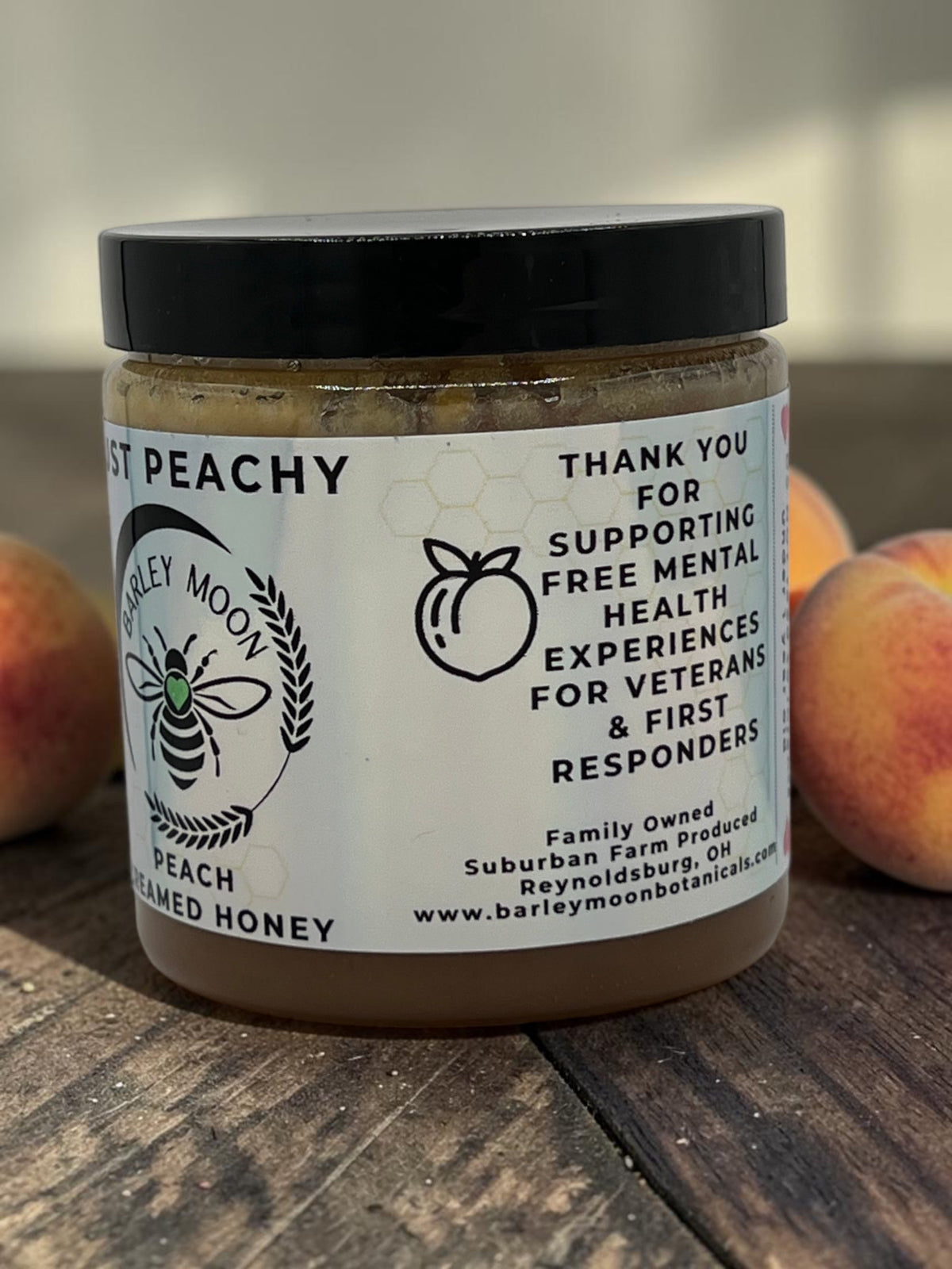 Barley Moon Botanicals Just Peachy creamed honey jar on a wooden surface with fresh peaches in the background.