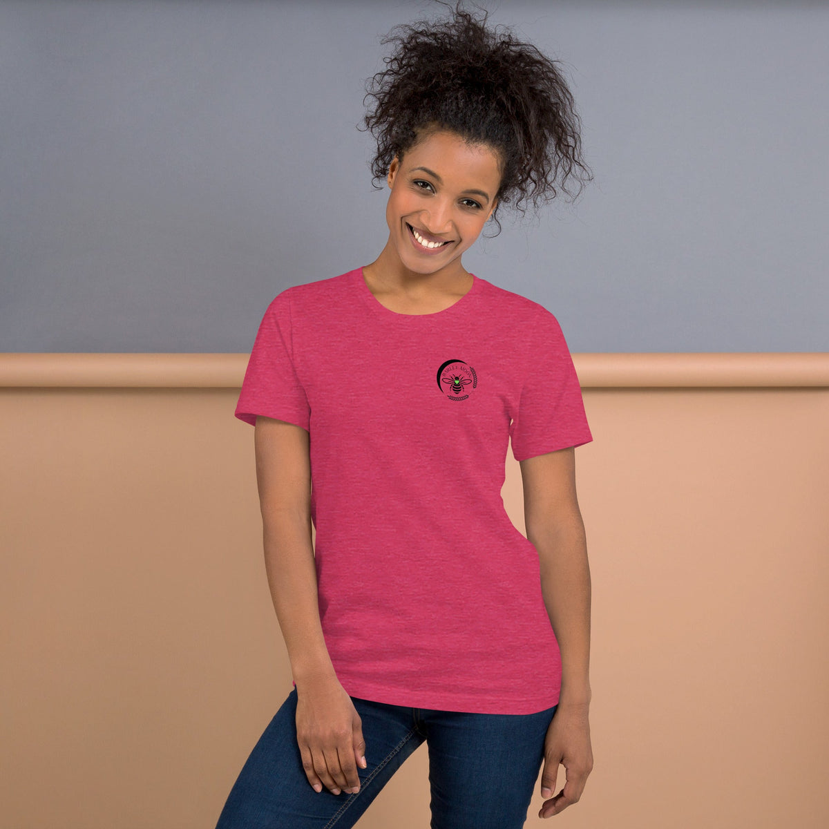 A smiling woman wearing a soft pink Barley Moon Botanicals t-shirt with a black bee and crescent moon logo on the chest.
