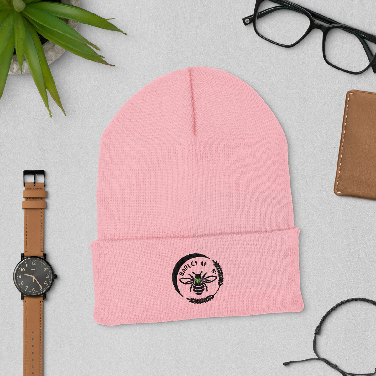 Barley Moon Botanicals pink cuffed beanie with a black bee and moon logo, displayed on a grey desk with accessories.