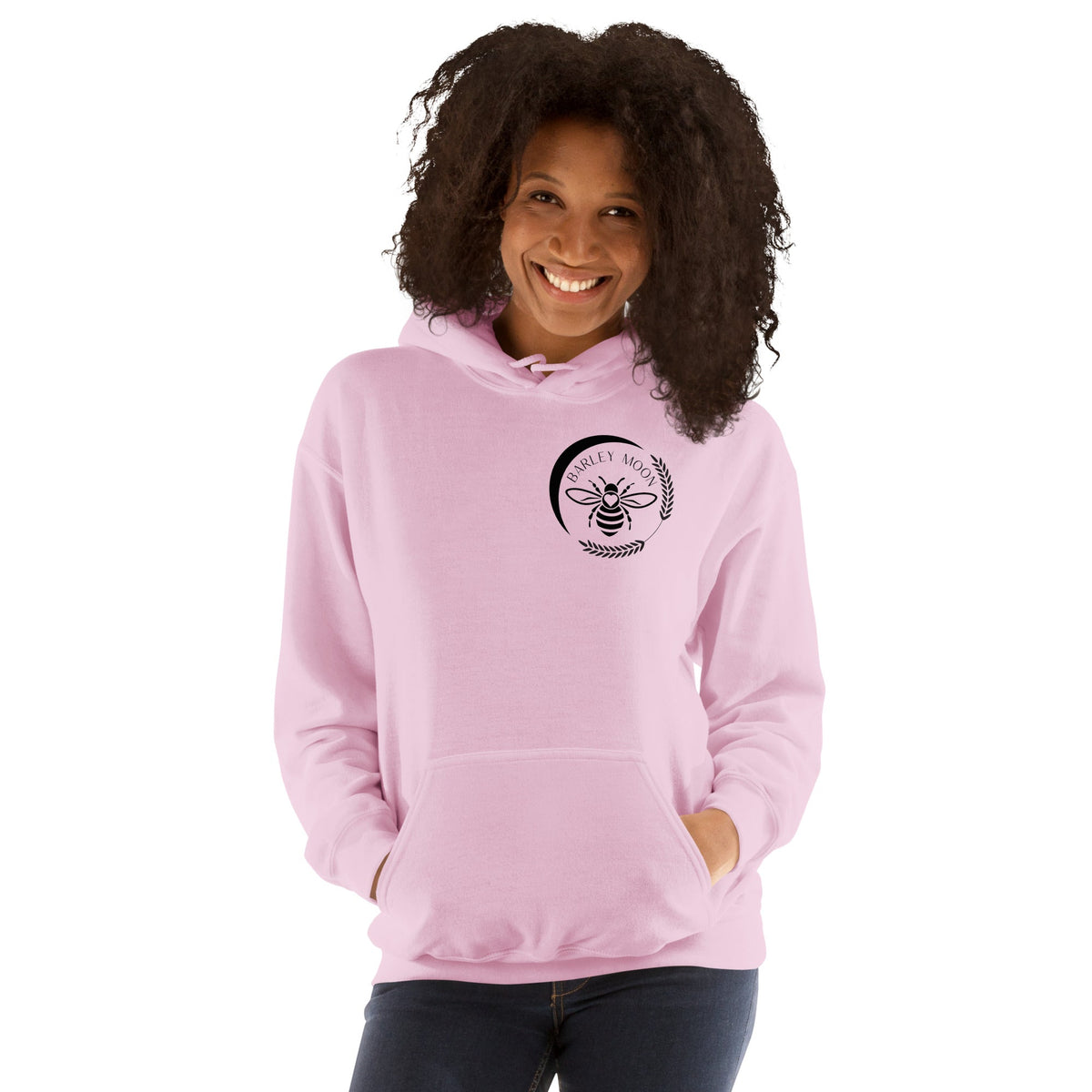 A woman wearing a light pink Barley Moon Botanicals unisex hoodie with a black bee and crescent moon logo on the chest.