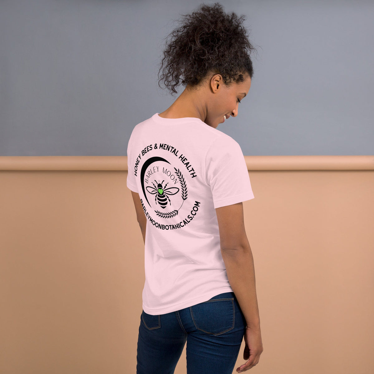 Woman wearing a light pink Barley Moon Botanicals t-shirt with a black bee and crescent moon logo on the back