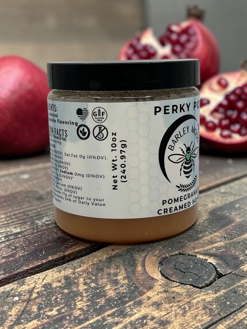 Barley Moon Botanicals Perky Pom pomegranate creamed honey jar on a rustic wooden table with fresh pomegranate halves.