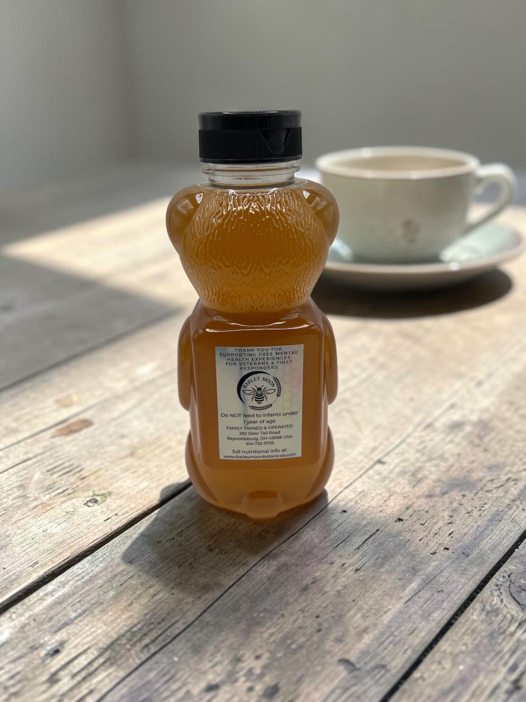Barley Moon Botanicals raw local honey in a classic bear-shaped bottle sitting on a rustic wooden table with a teacup