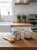 Barley Moon Botanicals raw honeycomb in a glass jar with a wooden spoon on a kitchen table next to a cup of coffee.