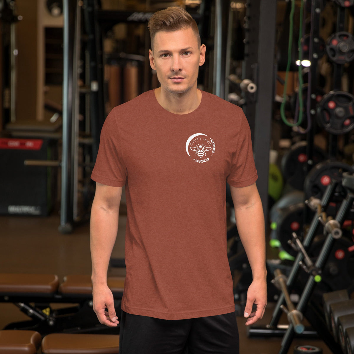 Man wearing a rust-colored Barley Moon Botanicals unisex cotton t-shirt with a white bee logo in a gym setting.