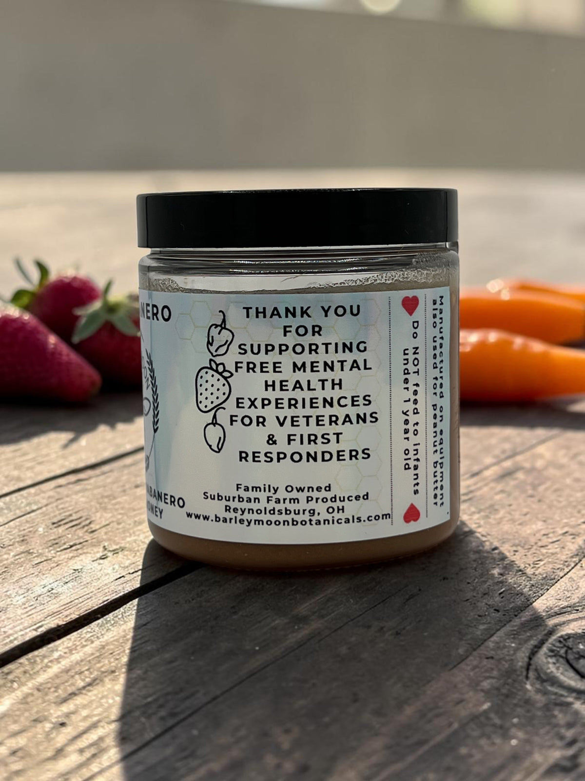 Barley Moon Botanicals Strawbanero creamed honey jar with fresh strawberries and habanero peppers on a wooden table.