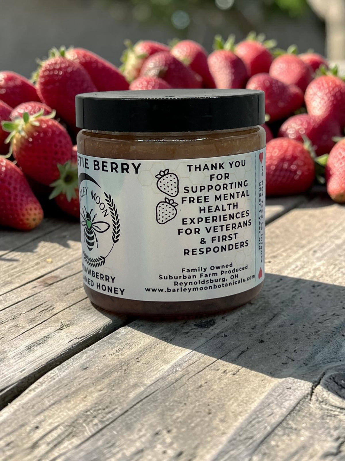 Barley Moon Botanicals Sweetie Berry strawberry creamed honey jar on a wooden surface with fresh strawberries behind.