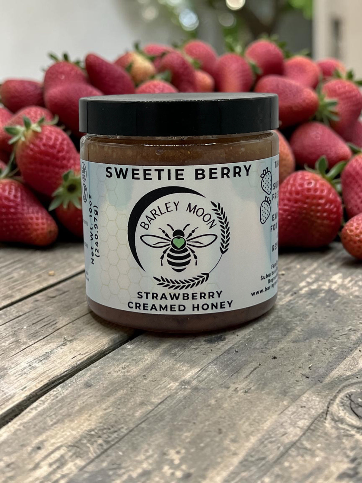 Barley Moon Botanicals Sweetie Berry strawberry creamed honey jar surrounded by fresh strawberries on a wooden table.