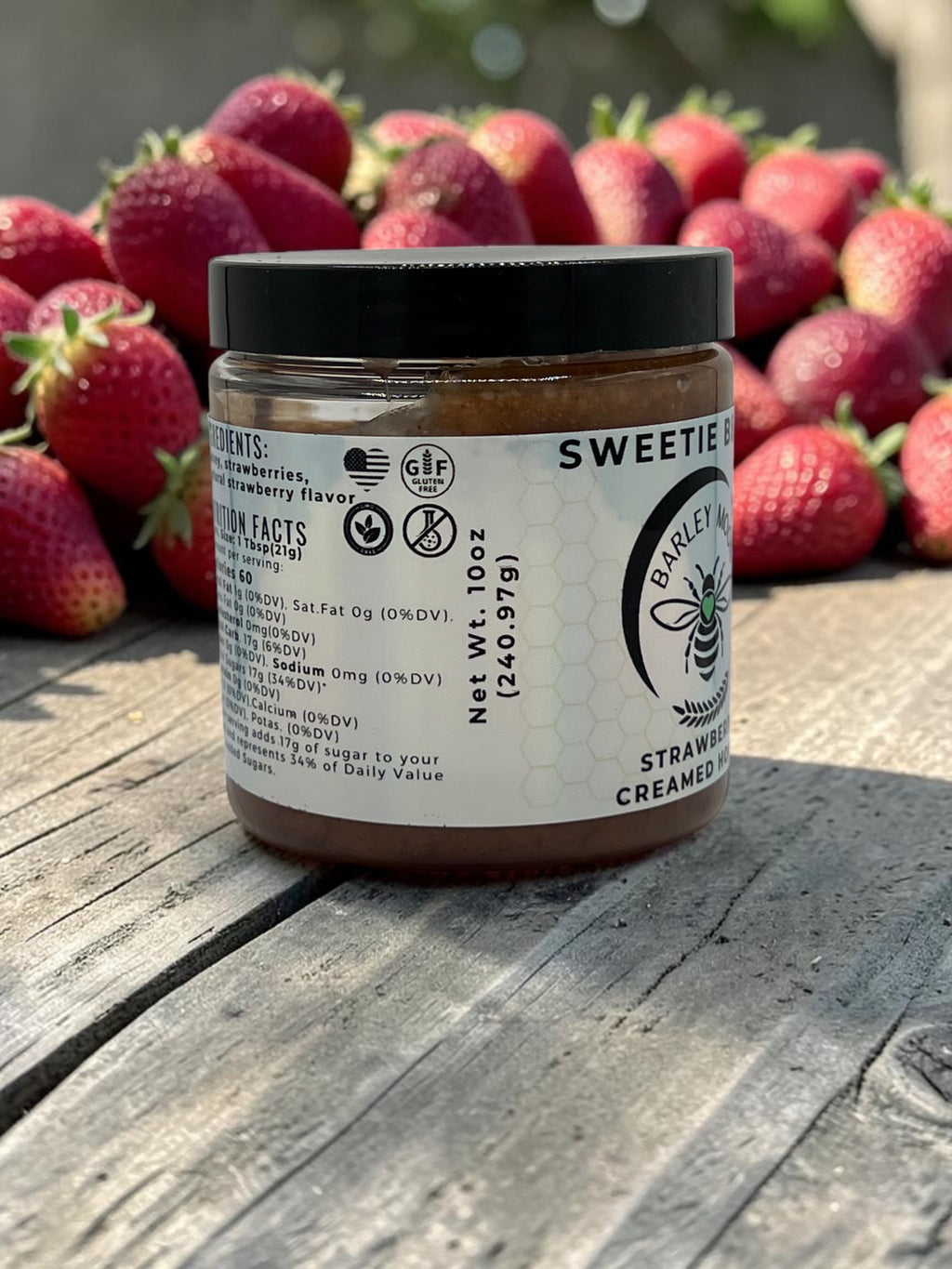 Barley Moon Botanicals Sweetie Berry strawberry creamed honey jar on a wooden surface with fresh strawberries behind.