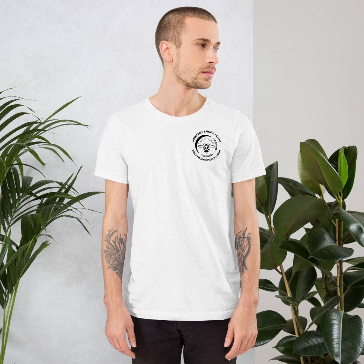 A man wearing a white Barley Moon Botanicals unisex t-shirt with a black bee logo on the chest in a plant-filled room.