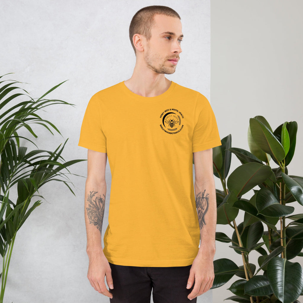 A man wearing a yellow Barley Moon Botanicals unisex t-shirt with a black bee logo on the chest, standing near plants.